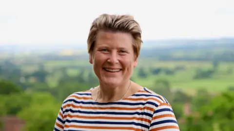 Michael Anderson Sarah Dyke wearing a blue and orange striped top and a silver necklace. She has short blonde hair and is smiling at the camera. In the background is a blurred landscape view looking over green fields and trees. 