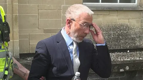 James Holder is escorted from Cirencester Magistrates' Court by a police officer. He is wearing a dark suit and grey tie
