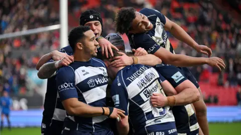 Getty Images A group of Bristol Bears rugby players huddle together as they celebrate a try against Newcastle. A player is leaping in the air as he joins the group. The match is at Ashton Gate and the players are wearing Bristol's traditional blue and white hooped shirts