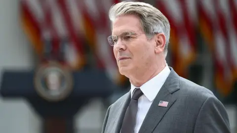 US Treasury Secretary Scott Bessent's head and shoulders in profile. He has grey hair and glasses. He is wearing a grey suit with a US flag lapel pin, a silver tie, and a white shirt. Behind him in soft focus are US flags and a podium.