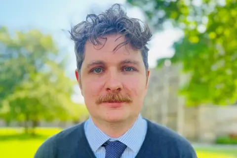Dan Cutts, Aberdeen co-chair of the UCU, a man with wavy dark brown hair, a moustache, and a blue shirt, tie, and jumper.