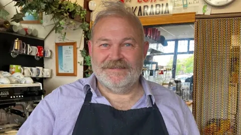 A man with short grey hair and a matching beard is standing in a cafe beside a large, black, Gaggia coffee machine. He is wearing a black apron over a lilac patterned shirt.