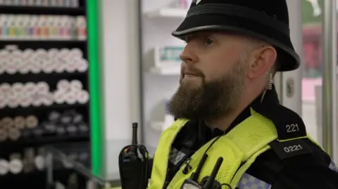 PC Bielby is standing in a shop. He is wearing police uniform including a yellow high vis vest with a body cam and radio attached as well as his police hat.