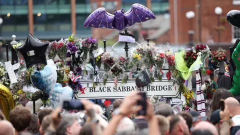 EPA A Black Sabbath Bridge sign with numerous tributes stuck to the railings, a bat ballon above them, and the heads of the crowd in the foreground