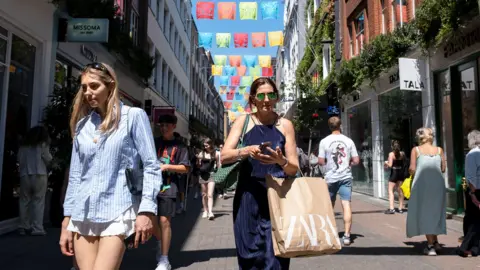 two shoppers on a busy shopping street in London walking towards the camera wearing summery clothing 
