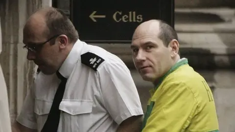 Getty Images Jack Whomes is being led through a door by a security officer wearing a white shirt and black tie. Whomes is wearing a yellow and green prison jumpsuit. He looks cautiously towards the camera.