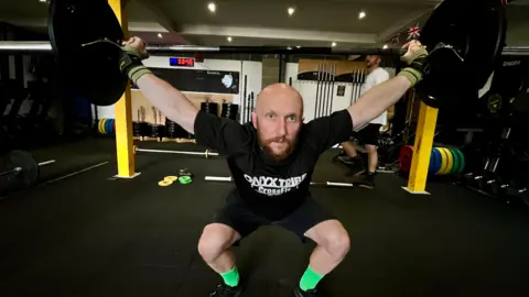 MARTIN GILES/BBC Lukas Koziel picks up weights on a bar above his head in a snatch. He has a brown beard and bright green socks. There are lots of weights in the background. He has grips on his hands and his arms are spread above him in a v shape. He has a black t shirt and shorts on.