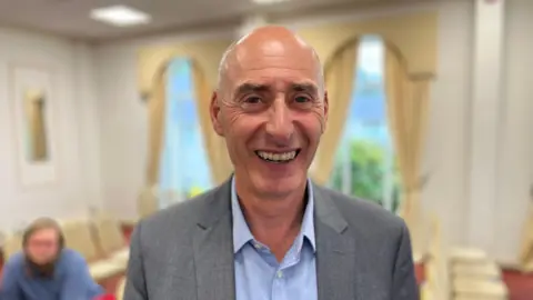 Housing minister Steve Williams is pictured in a head and shoulders shot. He is a balding man in a grey suit and blue shirt. He is smiling widely and is standing in a room filled with rows of chairs.