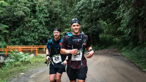 Jenny Scott Communications Two men running on a road with trees in the background. They are wearing numbers on their tops.