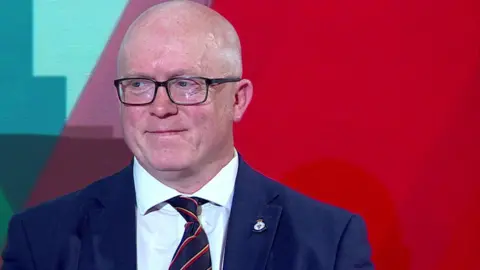 Head and shoulder shot of a balding Rob Potts. He is wearing glasses, a dark blue jacked with a small lapel badge, a white shirt and a dark blue tie with thin red and yellow stripes. The background to his left is red, with blue and purple patches on his right.