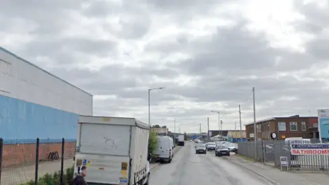 Vehicles are parked on either side of the road which runs through a partial industrial estate.