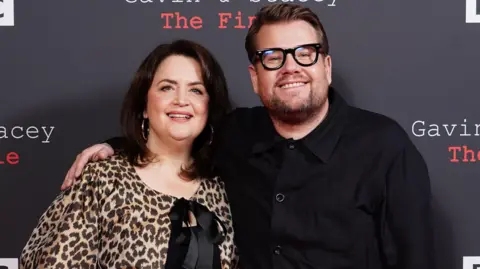 Ruth Jones is standing on the left, wearing a leopard print blouse tied with a black bow in the front. James Corden has his arm around Jones' shoulder, wearing a black shirt and thick black framed glasses