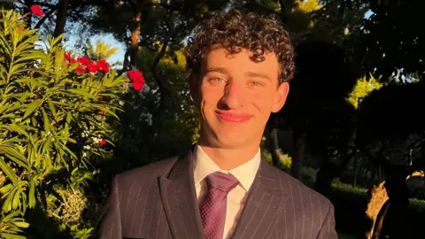 Daniel Daniel, who has dark curly hair and is wearing a purple suit stood outside with green bushes and red flowers in the background. 