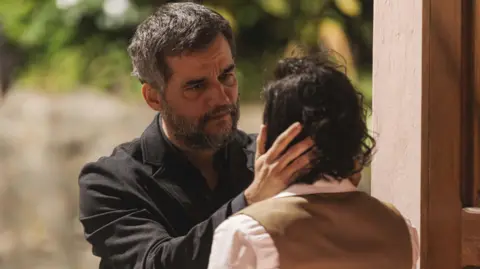 Caio Lirio Wagner Moura, in a dark suit with dark greying hair and beard, stares into the face and holds the head of a person with their back to us. It looks very serious.