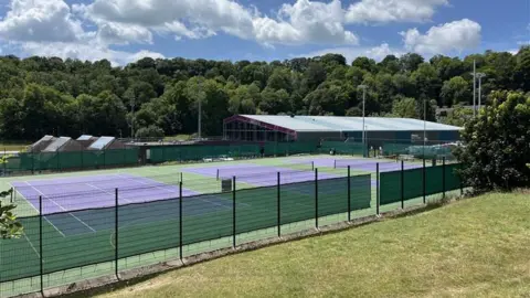 George Carden/BBC Three outdoor tennis courts at Withdean, which are purple with a green outside perimeter. They are surrounded by grass and trees. It is a sunny day.