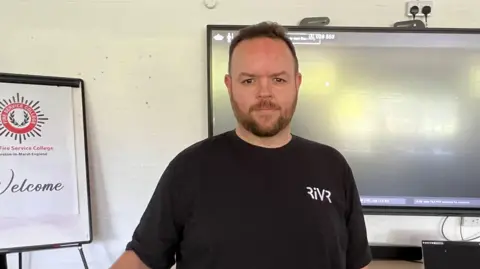 A mean wearing a black shirt with the RiVR logo standing in front of screen in a classroom.