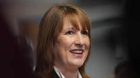 Chancellor Rachel Reeves smiles in a headshot. She wears a high-necked white shirt with a black jacket. Her light brown hair is work loose with a long fringe.