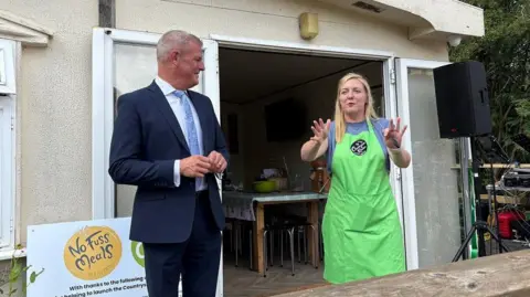 Martin Heath/BBC Stuart Andrew with short white hair, wearing a dark blue suit and light blue tie. Milly Fyfe has long blonde hair and is wearing a green apron over a blue T-shirt. She has her hands outstretched while talking. They are standing outside a single-storey wooden classroom with doors open revealing a table inside.