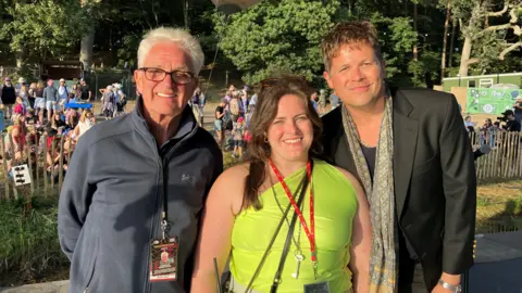Jon Wright/BBC A man, a woman and a man stand in a line smiling at the camera. They are on a stage at a music festival with crowds in the background.