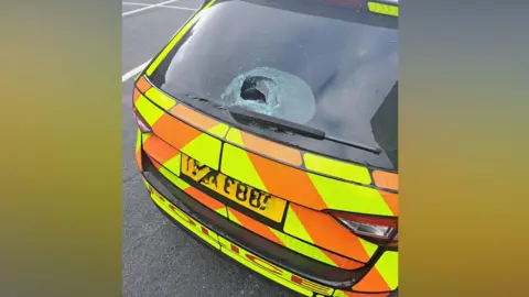 An orange and yellow marked PSNI car. The photo is taken from behind the car and the window has been damaged, with a hole in it. 