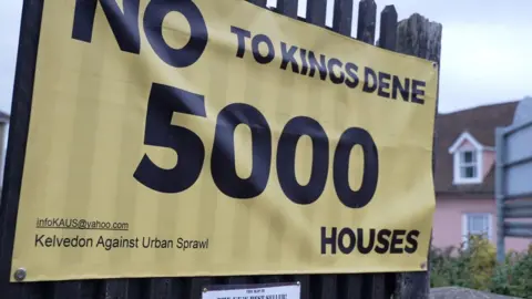 John Fairhall/BBC A large yellow sign says No To Kings Dene 5000 Houses in black print. The sign is pinned to a fence post. 