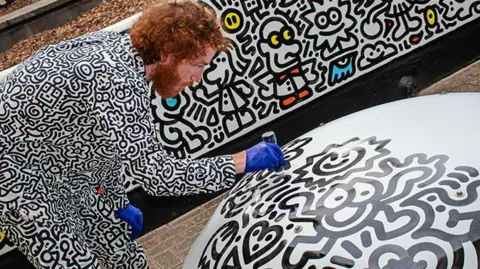 A man with ginger hair and beard wearing a black and white doodle suit drawing on a slab of concrete 