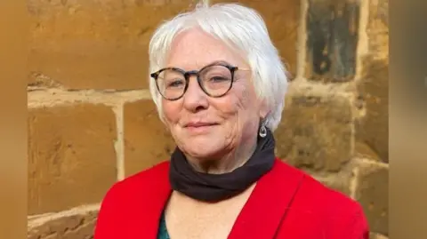 Danielle Stone with light-coloured hair and red top with a brick wall behind her