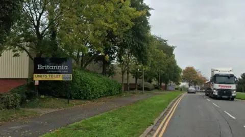 A road with a double yellow line along the side of it. There is also a footpath running alongside and a grass verge along with a row of trees. There is a large truck heading towards the direction of the photographer.