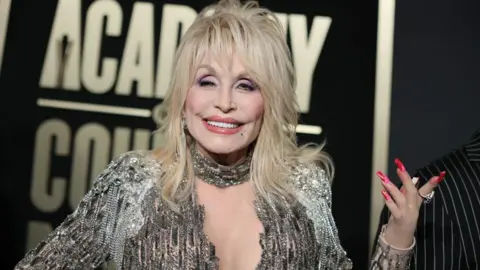 Getty Images Dolly Parton is stood in a silver dress, smiling.
