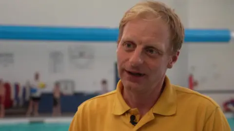 A man in a yellow polo shirt with blonde hair standing in front of an indoor swimming pool with a few swimmers in the background.