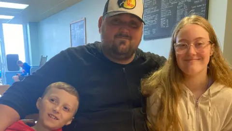 Supplied Luke is wearing a cap and has his arms around both of his children. His daughter is wearing glasses and has long blonde hair and his son has a red jumper.