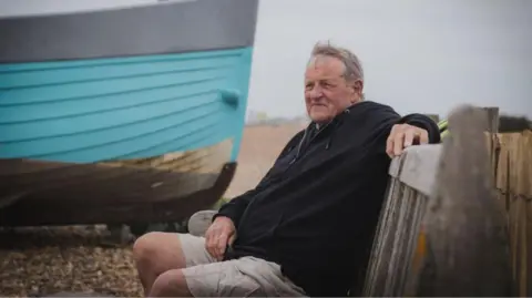 Urszula Soltys A man wearing a black hooded top and grey shorts. He is sat on a bench with a blue boat on a pebbled beach behind him.