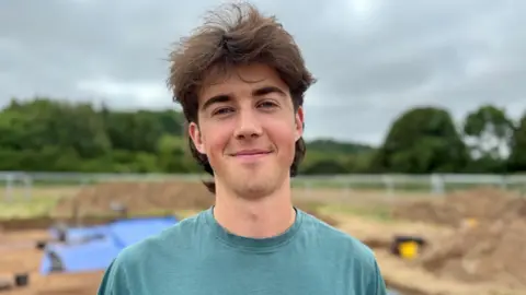 Cardiff University Student Dan Quelly, in his teens, stands on the archaeology site at Trelai Park. The background is blurred but you can see the excavatin site. He wears a green t-shirt and smiles at the camera. He has a brown mullet and blue eyes.