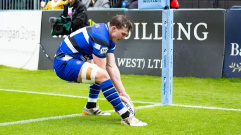 Ted Hill, wearing a blue shirt with black and white stripes, placing the ball over the try line to score near the corner flag during a game between Bath and Newcastle.