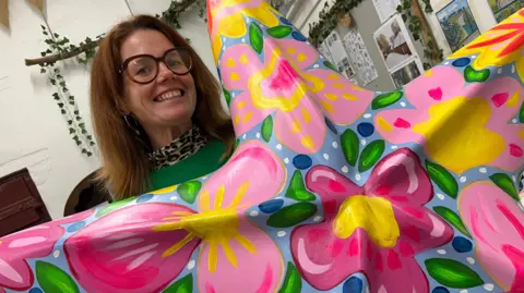 A woman smiles as she holds a large starfish sculpture painted in a bold pink and yellow floral design. She has red brown hair, large tortoiseshell glasses and is wearing a leopard-print polo-neck beneath a jade green jumper.