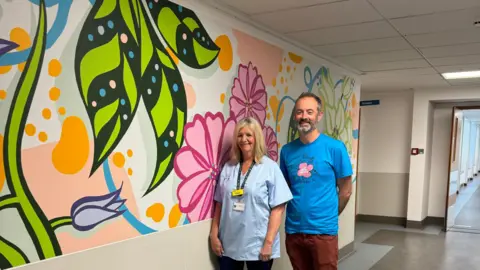 A man and woman stand in front of the mural at Princess Elizabeth Hospital.