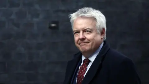 Getty Images Carwyn Jones yang berambut abu-abu mengenakan dasi bermotif merah, kemeja bergaris vertikal, dan jas berwarna gelap. Di belakangnya ada tembok bata abu-abu