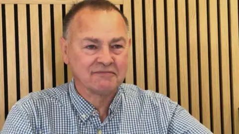 An image of Dr Matt Cole taken in front of a slatted wooden wall. He is wearing a checked blue and white shirt and is smiling.