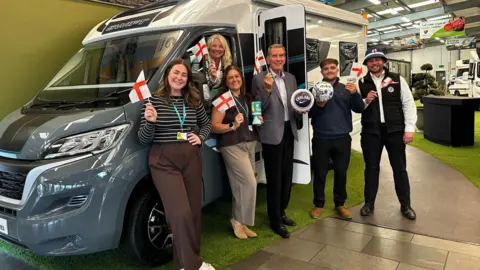 Salop Leisure A group of five people standing in front of a white and grey motor home that is inside a showroom. Another person is in the vehicle and peering out of the window. They are holding small England flags and an older man in the middle is holding a football. The motor home is parked on a piece of fake grass. They are all smiling. 