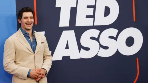 Getty Images Phil Dunster, wearing a cream jacket and blue shirt, standing in front of a sign for TV programme Ted Lasso. He has dark hair and is smiling.