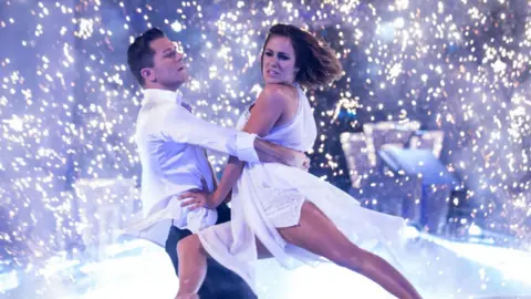 Guy Levy/BBC Pasha Kovalev, in a white shirt and dark trousers, and Caroline Flack, in a white floaty dress, mid-dance, Caroline lifted by Pasha as bright white lights create a backdrop