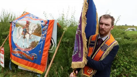 Reuters A man is stood in front of a hedge. He is wearing a dark suit and an orange sash around his shoulders. In his right hand is a flag with golden tassels. In the hedge to his right is a flag of King William on a horse.