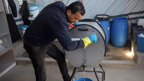 A man is turning a hand-cranked washing machine, which is a silver metal barrel with a tap at the bottom of it. Some water is coming out of the tap into a blue bucket. The man has dark hair and is wearing a navy fleece and black jeans. He is wearing blue and yellow rubber gloves. There are other barrels and buckets behind him, some with fabric in them.