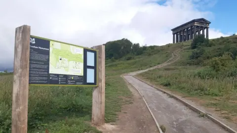 LDRS A swerving path leading up to Penshaw Monument, which resembles a Greek temple with stone pillars. There is a National Trust sign at the bottom of the hill with a map and information about the site.