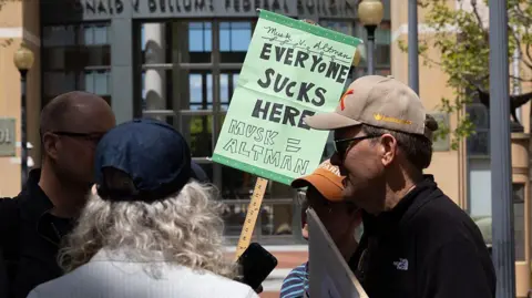 Getty A group of four people gathered outside a building. One is holding a green sign which reads "Musk v Altman - Everyone Sucks Here - Musk = Altman".
