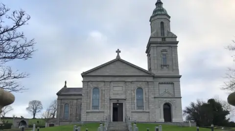 St John the Evangelist Church, Ballymaghery - a large grey building with a bell tower standing on a hill in front of steps and gardens. A graveyard and trees are in the background.