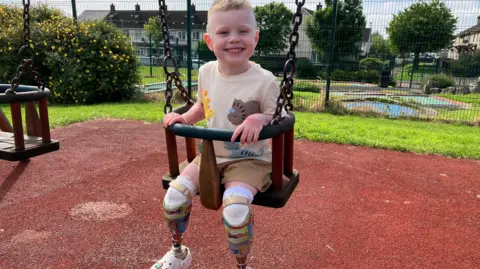 Dáibhin McConnell-Mailey sitting on a children's swing in a playpark on a sunny day. He is smiling at the camera and wearing a beige t-shirt adorned by yellow and brown dinosaur motifs and brown shorts. He has two prosthetic legs and is wearing white croc shoes. 