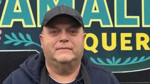 Darin Alberry Man standing outside food truck with blue hoodie and black apron on.