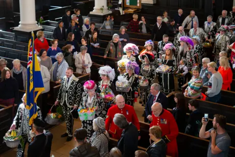 Getty Images A line of pearly kings and queens dressed in black suits with white pearls walk down the aisle of St Martin-in-the-Fields with people standing in the pews