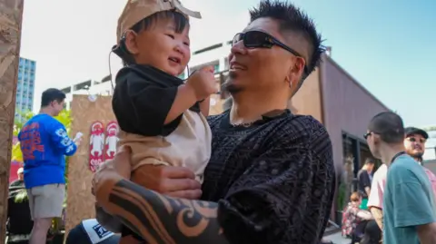 A man with a tattoo on his arm is smiling while looking at a smiling toddler he is carrying.